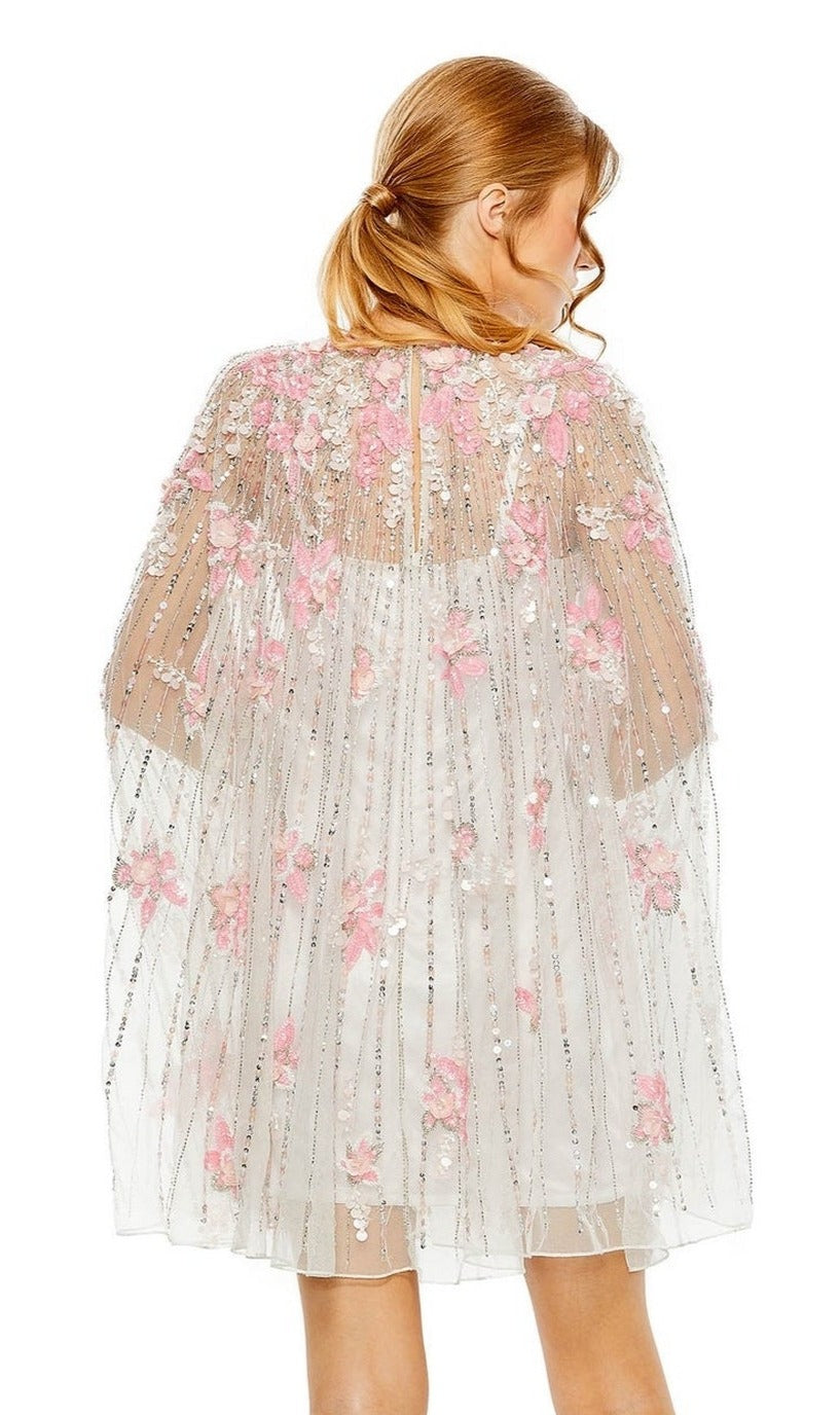 A woman with light brown hair faces away, wearing the Beaded-Cape Short Hoco Dress by Mac Duggal 5917—a sheer cape with pink and white floral appliqués and vertical beaded lines, layered over a short dress.